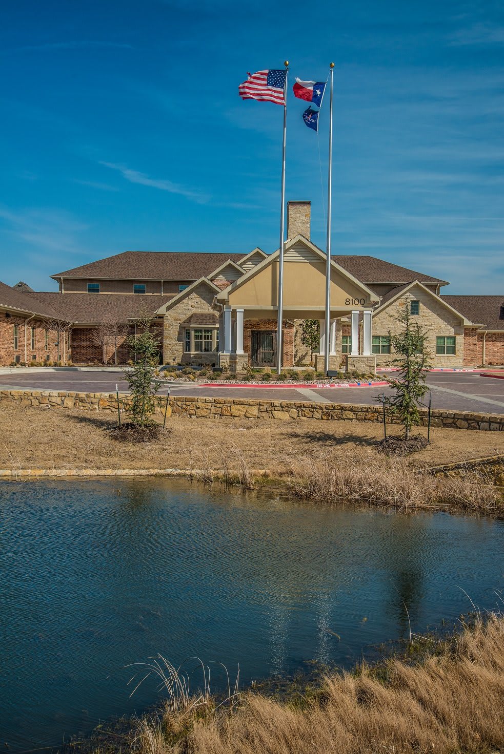 A large building with a pond in the foreground, featuring an entrance with flags and surrounded by landscaping under a blue sky.
