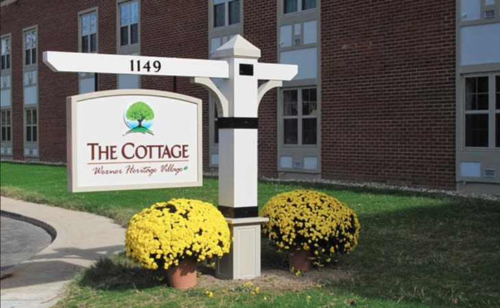 A sign for 'The Cottage' in front of a brick building with yellow flowers.
