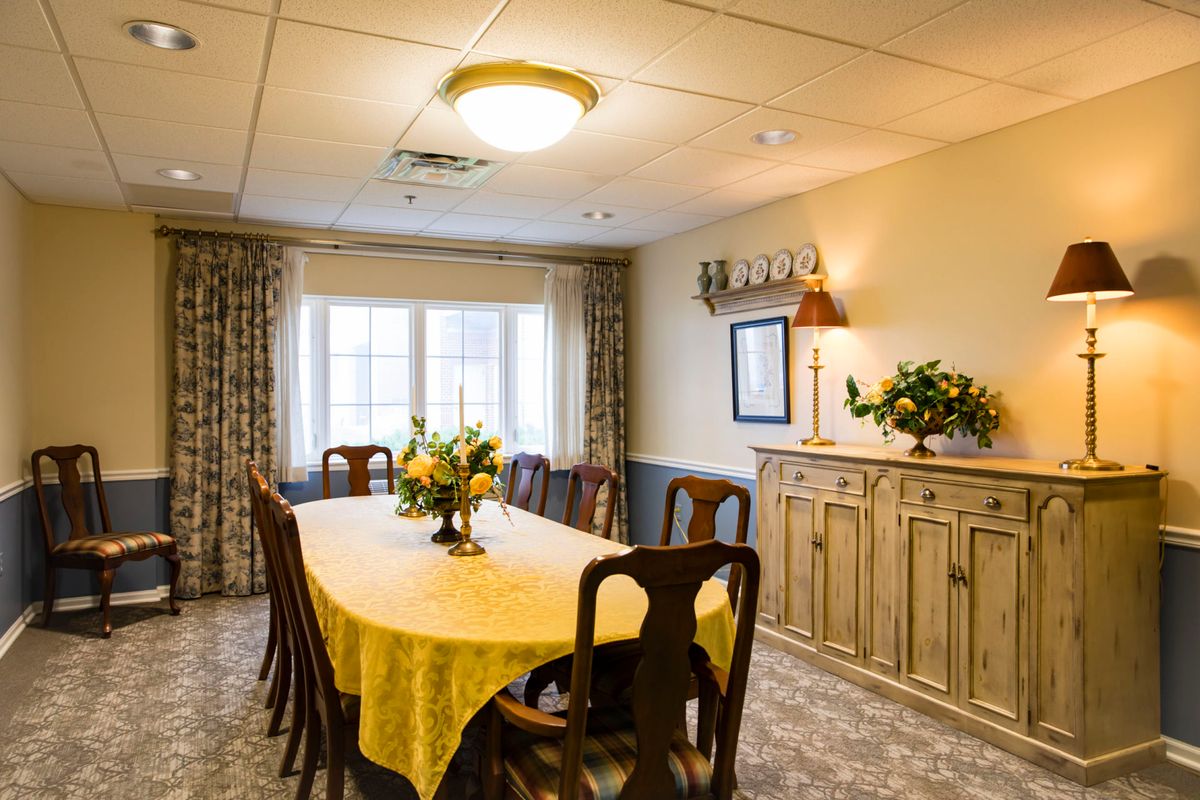 A cozy dining room with a long table covered in a yellow tablecloth, surrounded by wooden chairs. The room features a window with curtains, floral decorations, and a wooden cabinet.