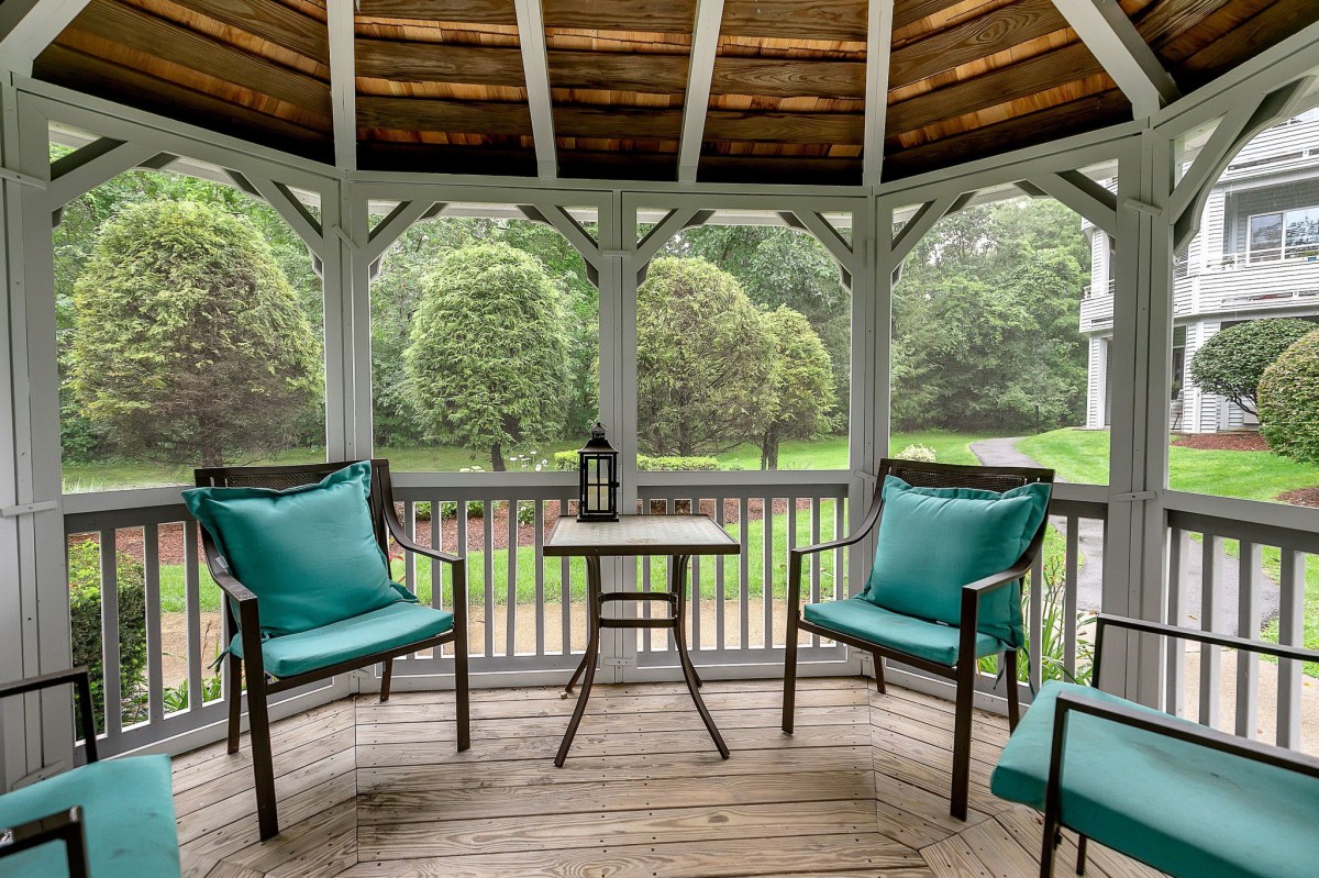A cozy gazebo with comfortable seating and a small table, surrounded by lush greenery.
