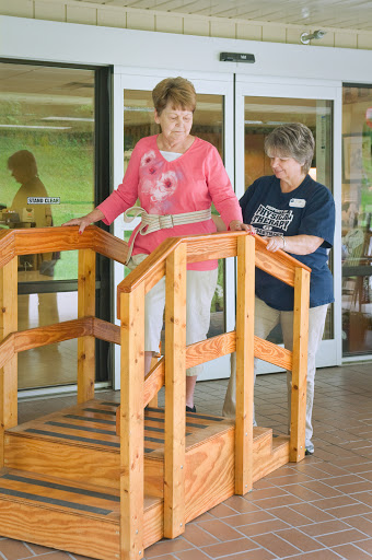 A caregiver assists an elderly individual as they practice walking on a wooden therapy ramp.