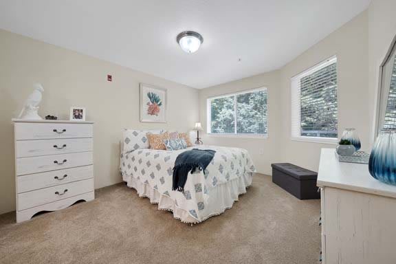A cozy bedroom featuring a bed with a patterned cover, surrounded by furniture and large windows providing natural light.