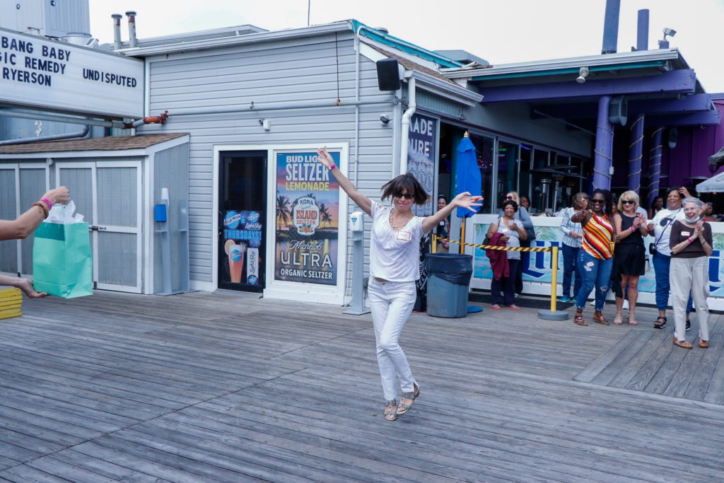 A person dancing energetically on a boardwalk in front of a shop, while others watch.