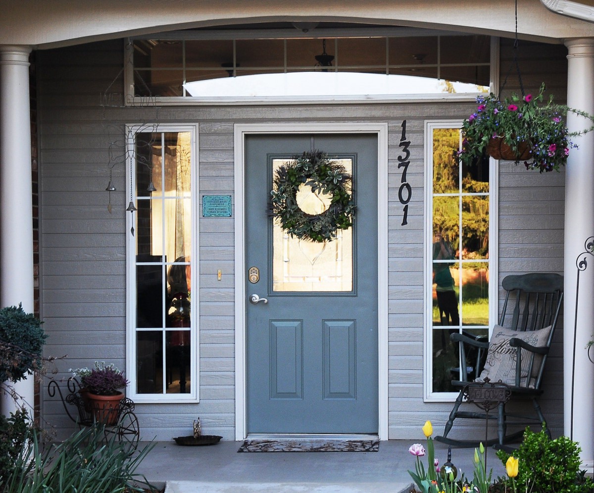 A cozy front porch with a gray door adorned with a wreath, flanked by windows and surrounded by plants.