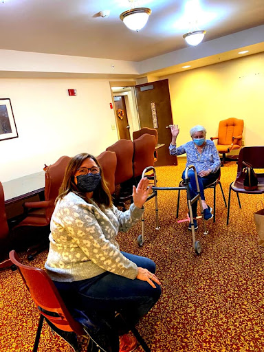 Two individuals engage in friendly interaction in a community room, both wearing masks.