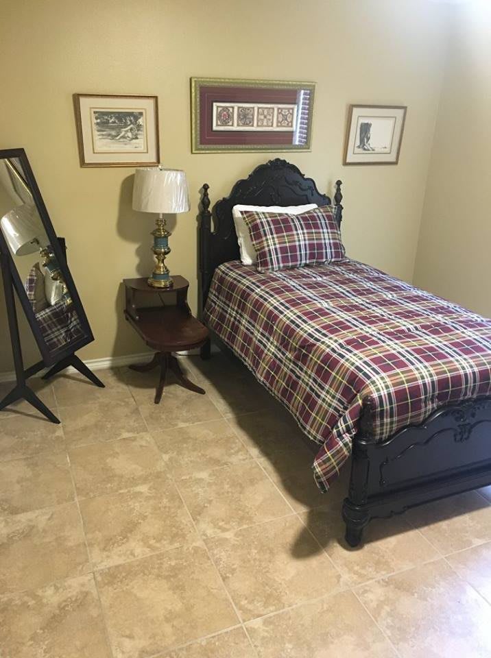 A cozy bedroom featuring a single bed with a plaid comforter, decorative frames on the walls, and a standing mirror beside a bedside table with a lamp.
