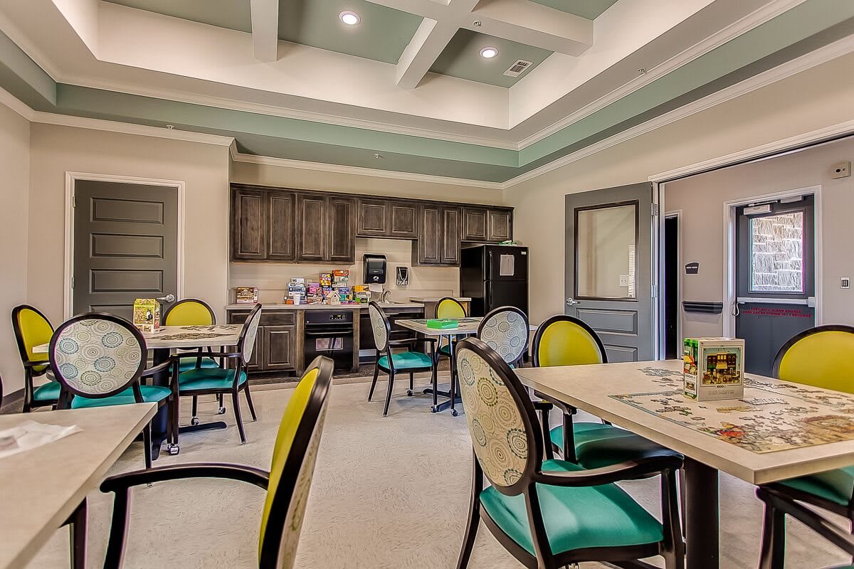 A well-lit dining area with colorful chairs and tables, featuring a kitchenette in the background.