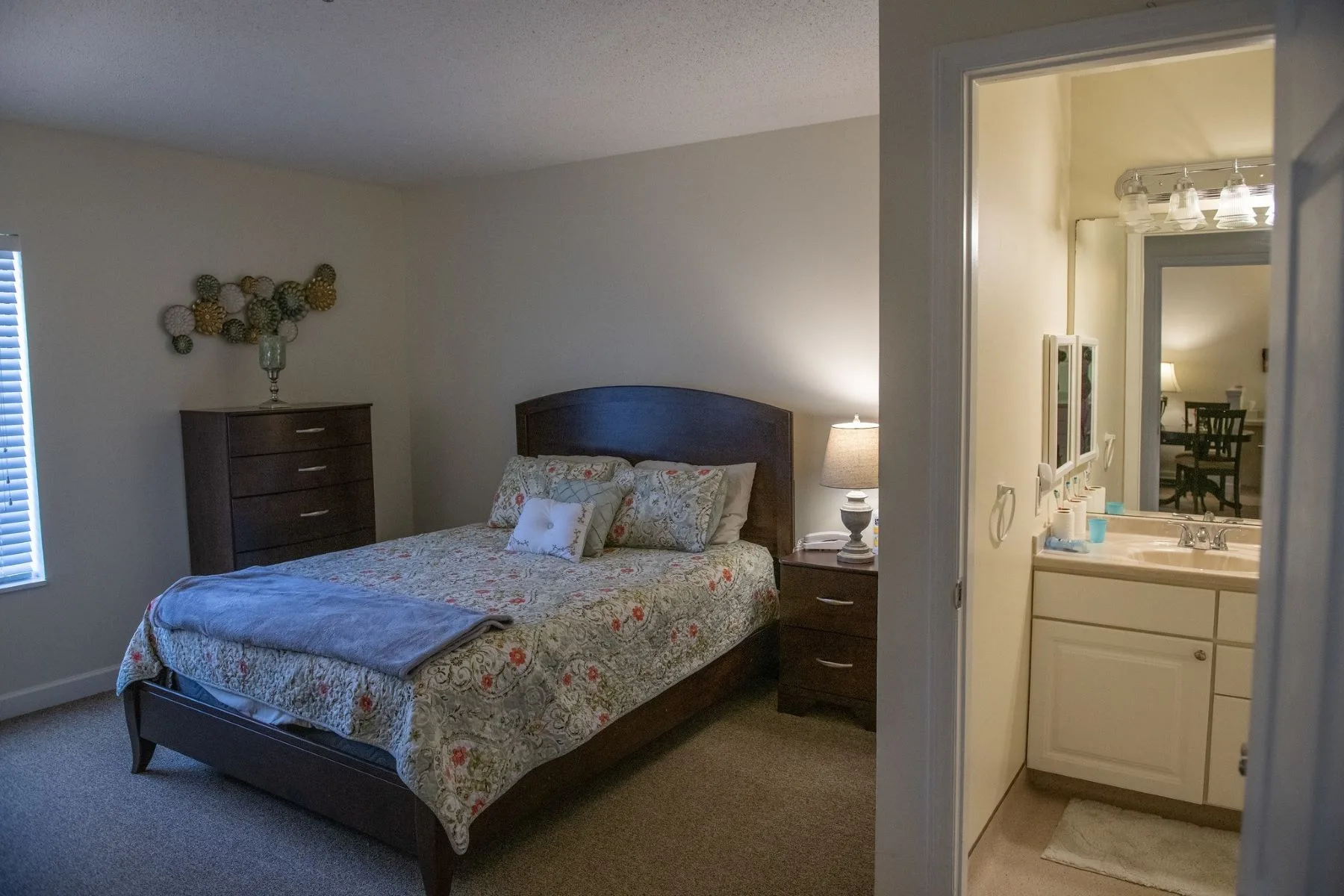 A cozy bedroom interior featuring a neatly made bed, nightstand with a lamp, and an open door leading to a bathroom.