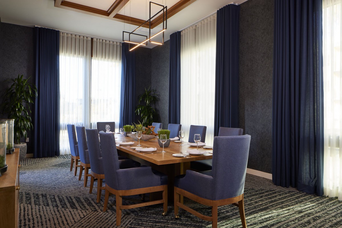 A modern and elegant dining or conference room featuring a large wooden table surrounded by upholstered chairs, with soft lighting and stylish curtains.