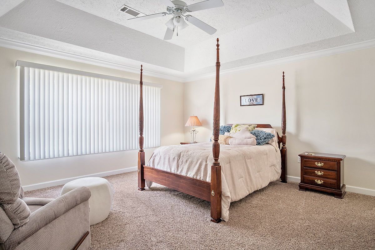 A cozy bedroom with a four-poster bed, carpeted floor, and natural light coming through the window blinds.
