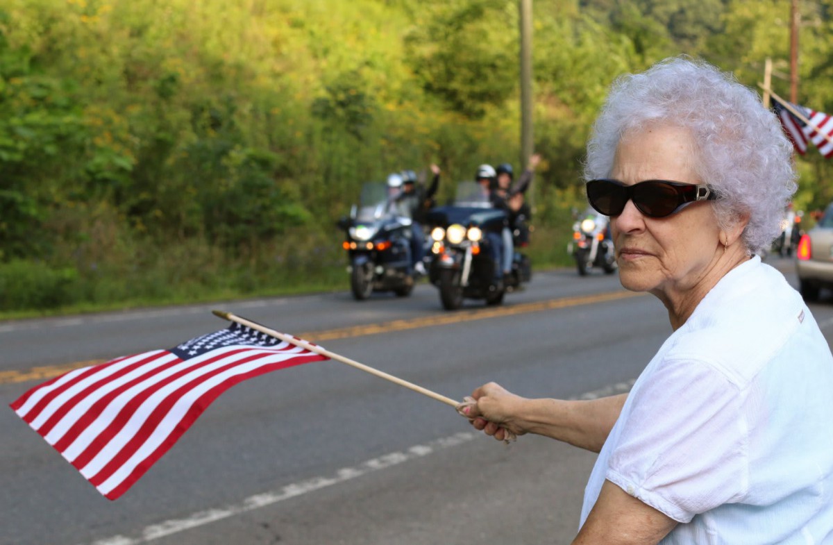 An elderly woman waves an American flag by the roadside as motorcycles pass by.