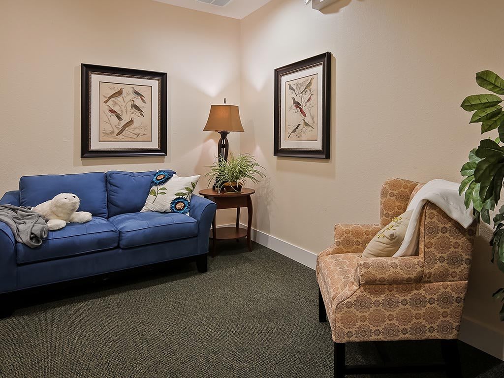 A cozy living room with a blue sofa and patterned chair, accented by artwork and a lamp.
