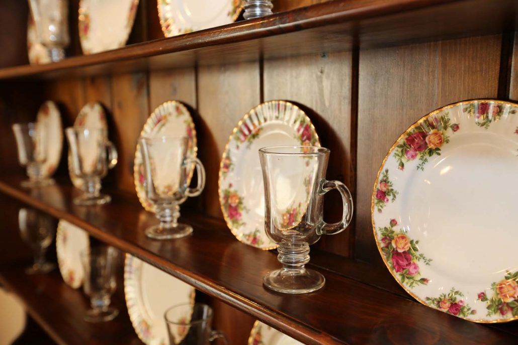 A collection of vintage floral china plates and glass mugs displayed on a wooden shelf.