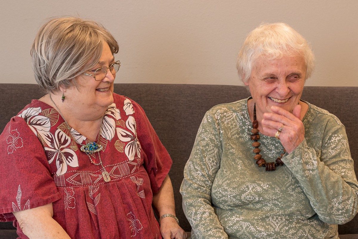 Two elderly women are sitting together on a couch, sharing a laugh.