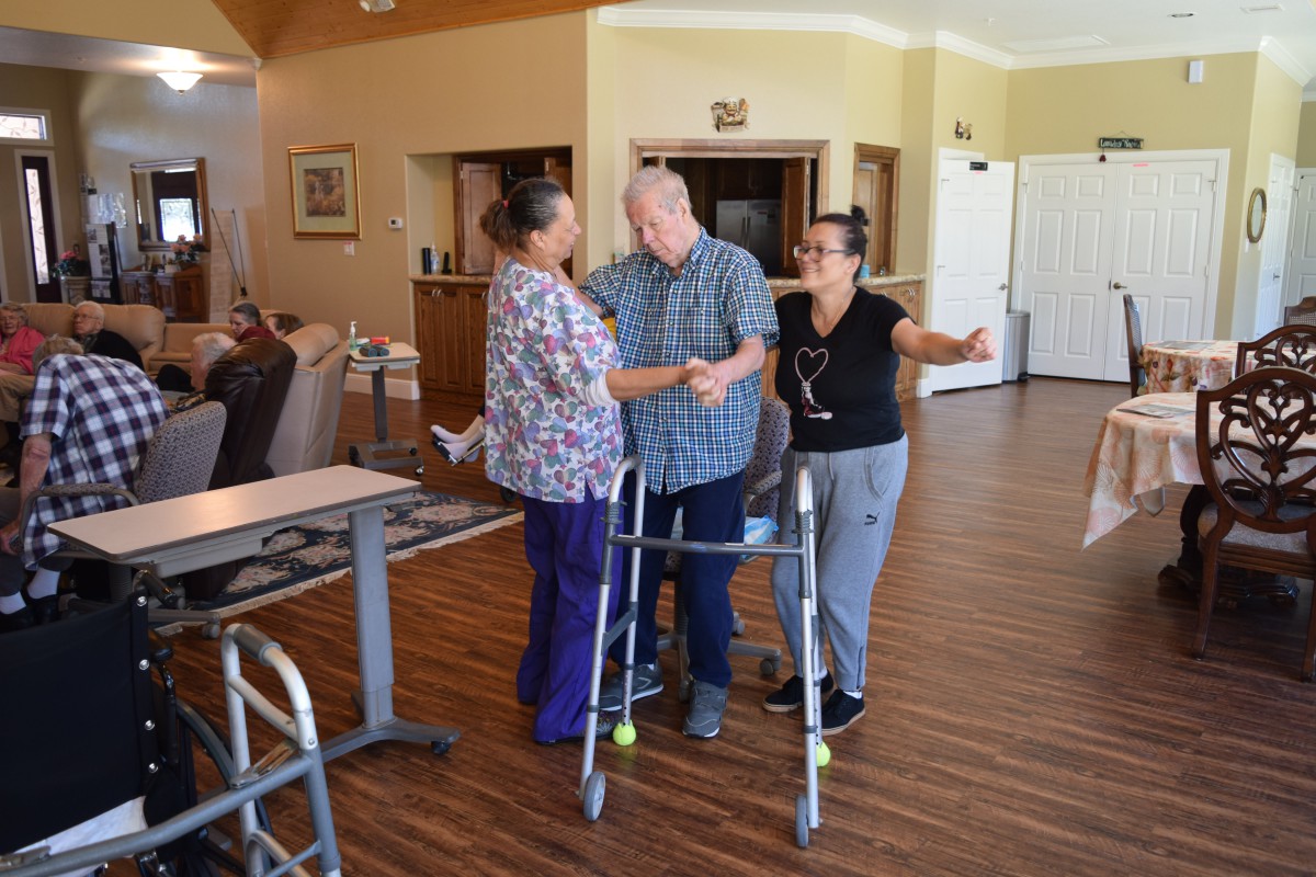 Caregivers assist an elderly man using a walker in a nursing home setting.