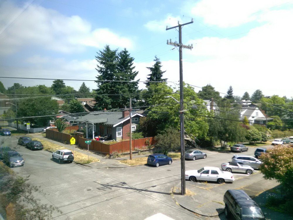 A suburban neighborhood street with parked cars and houses surrounded by trees.