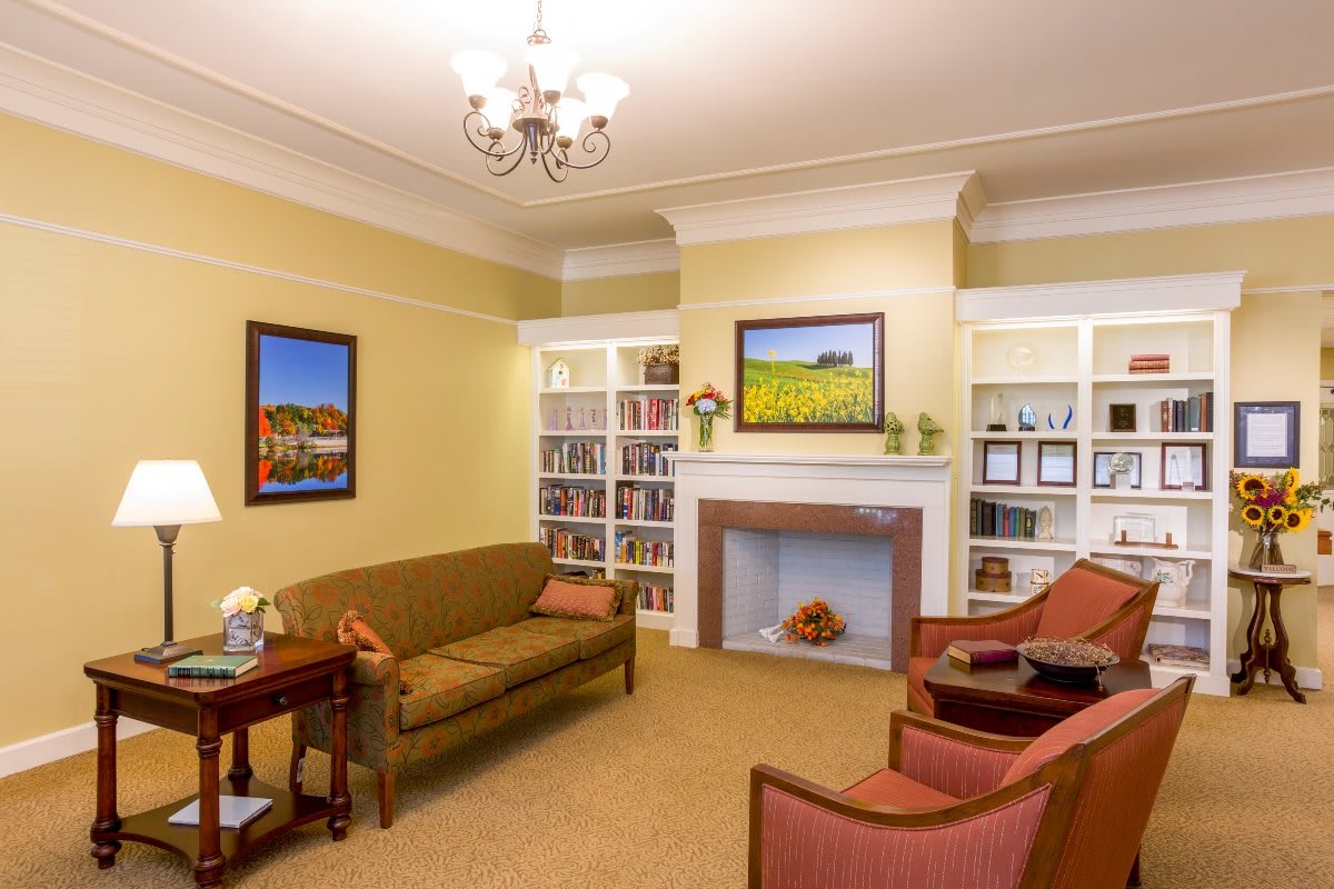 A cozy living room with traditional decor, featuring a sofa and armchairs around a fireplace, surrounded by bookshelves and decorated with framed artwork.