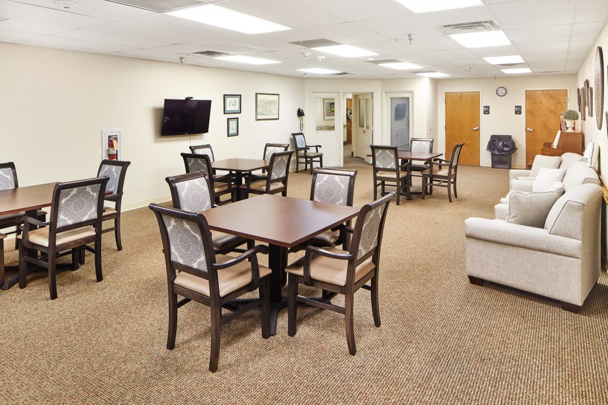 A well-lit dining area with multiple tables and chairs arranged on a carpeted floor, featuring a wall-mounted television.
