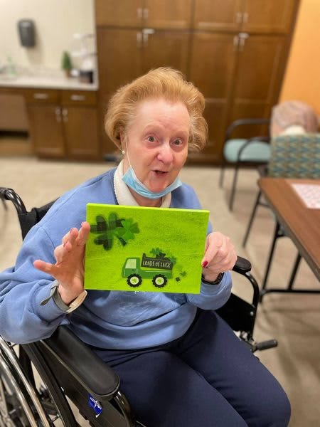An elderly woman in a wheelchair proudly displays her colorful painting indoors.