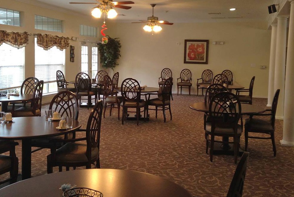An empty dining room with neatly arranged chairs and tables, featuring overhead lighting and carpeted flooring.