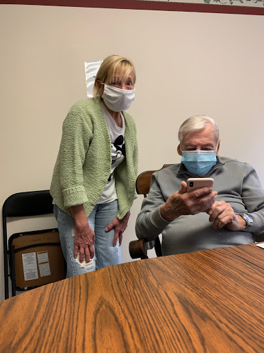A woman and an elderly man are interacting indoors, both wearing masks. The man is focused on his smartphone while the woman stands beside him.