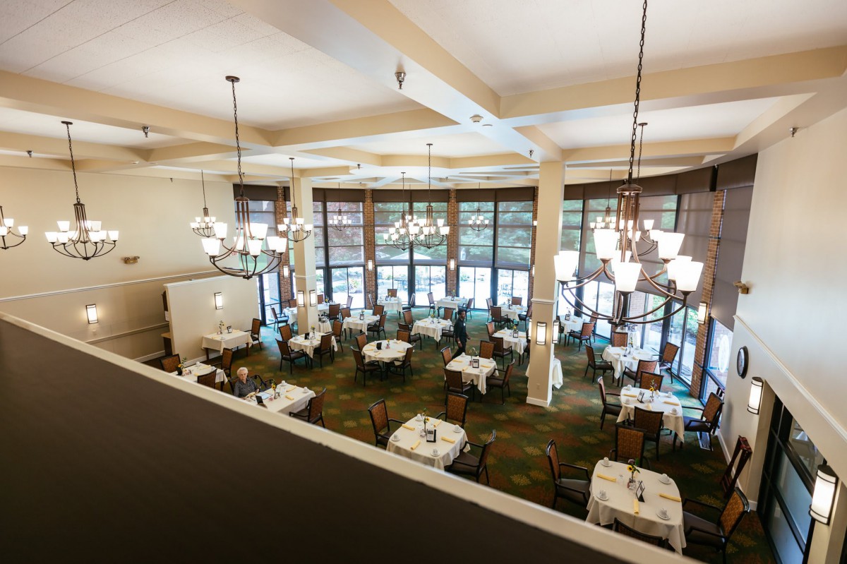 A spacious restaurant dining room with elegantly set tables and large windows, viewed from above.