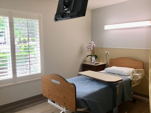 A well-lit hospital patient room featuring a bed, bedside table, and a wall-mounted TV.