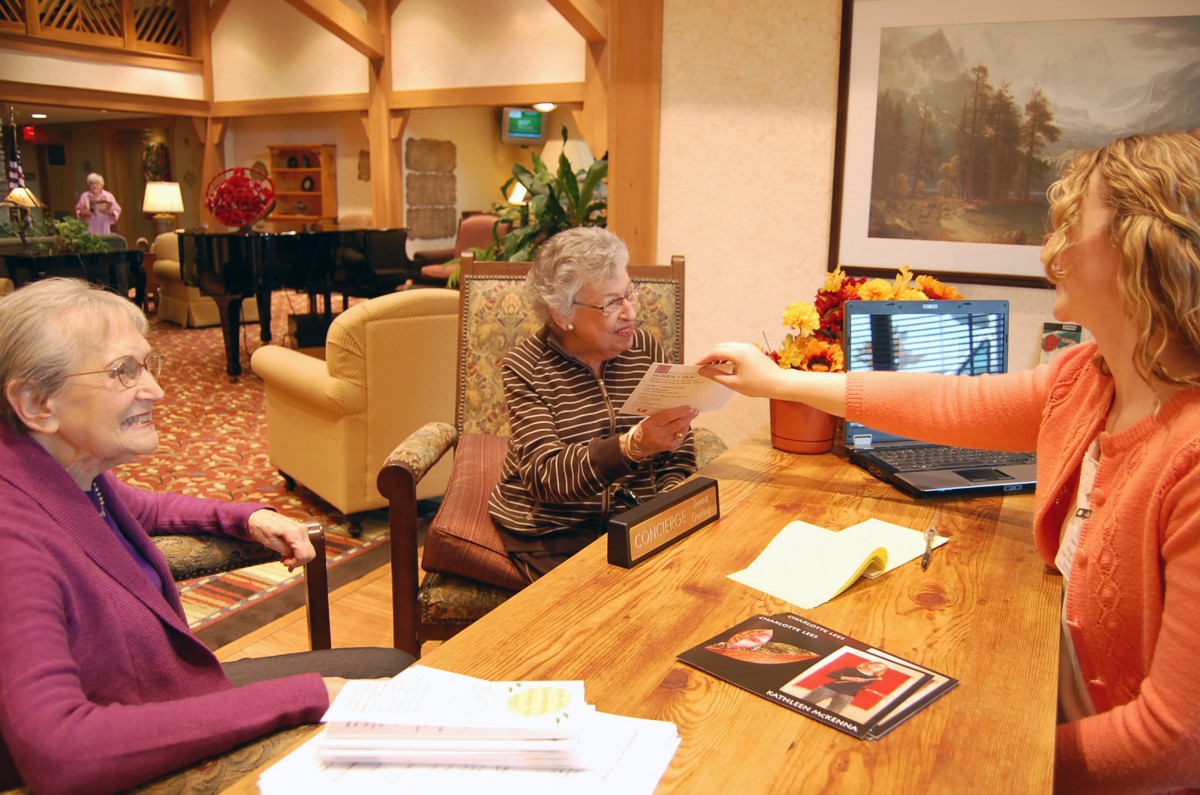 An elderly woman sitting at a table in an assisted living facility, engaging in conversation and exchanging papers.