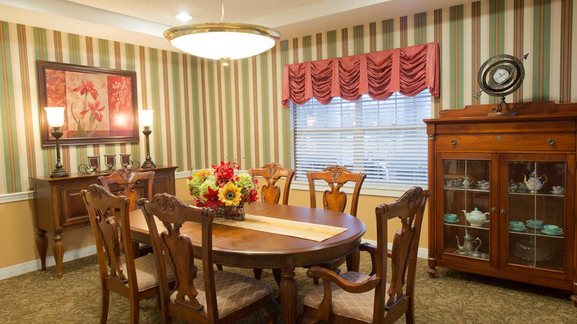 A cozy dining room featuring a wooden table with floral centerpiece, surrounded by matching chairs, set against striped wallpaper and illuminated by a chandelier.