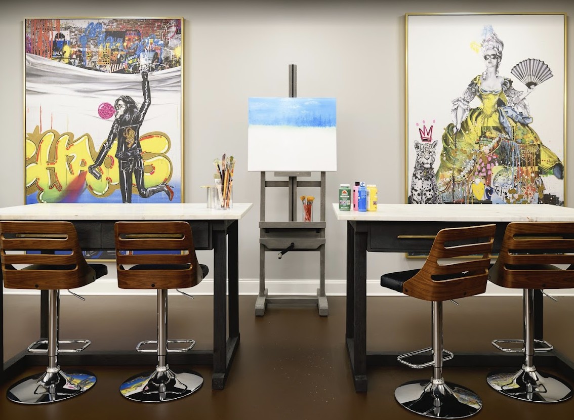 An art studio with tables, chairs, and an easel set up for painting. Colorful artwork is displayed on the walls.