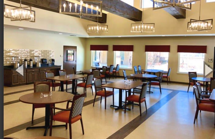 A spacious dining area with neatly arranged tables and comfortable chairs, illuminated by elegant chandeliers and natural light from large windows.