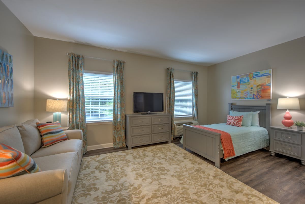A cozy bedroom with a comfortable bed, sofa, and television, featuring modern decor and ample natural light.