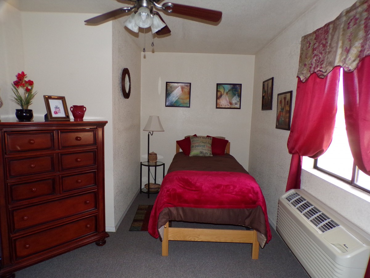 A cozy bedroom with a single bed featuring red bedding, a wooden dresser, a lamp, and artwork on the walls.