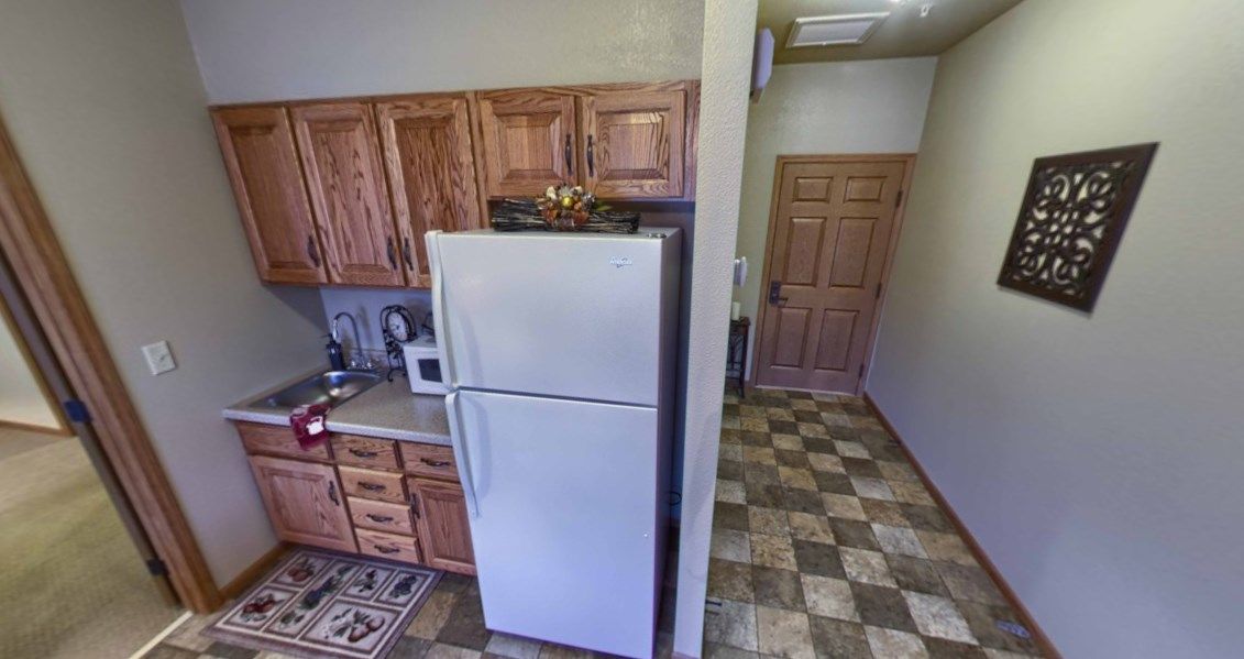 A small kitchen with wooden cabinets, a white refrigerator, and a microwave, leading into a tiled hallway.