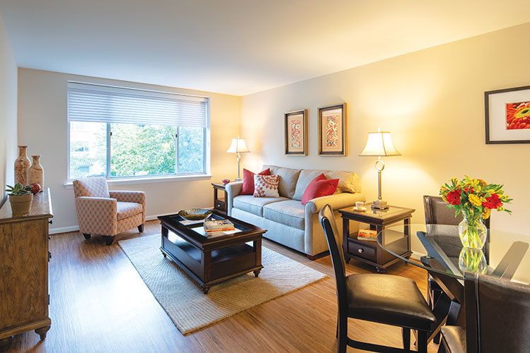 A cozy living room with a sofa, armchair, and coffee table, softly lit by lamps near a large window.