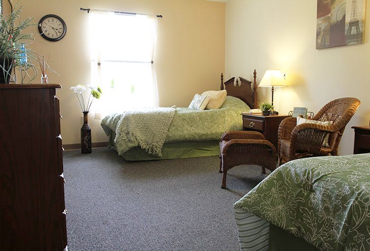 A cozy bedroom featuring twin beds with matching green bedding, illuminated by a table lamp and natural light from the window.