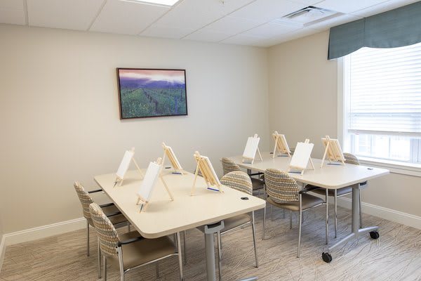 A well-lit classroom with tables and chairs arranged neatly, equipped with easels for painting activities.