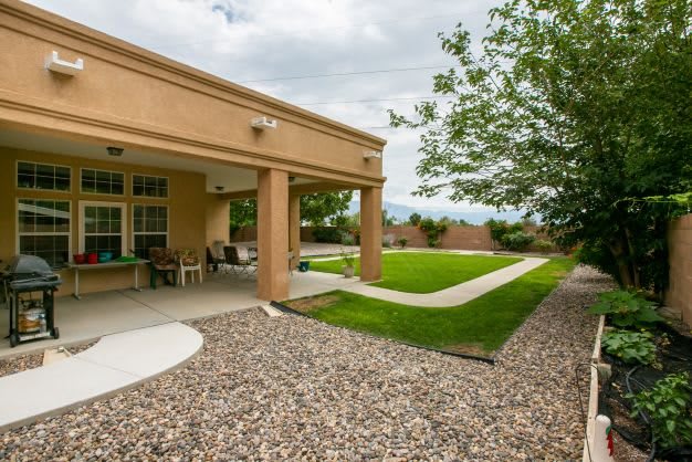 A modern house with a covered patio overlooking a green lawn and trees in the backyard.