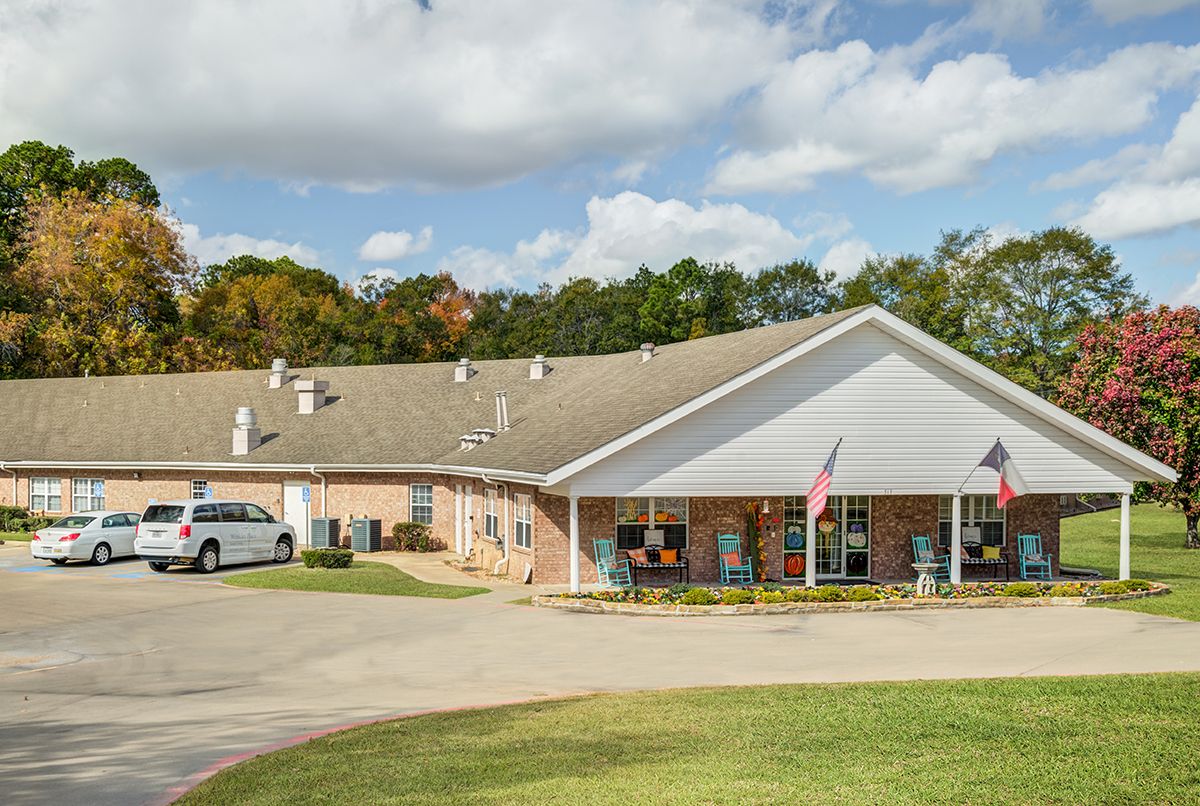 A one-story commercial building with a parking area, surrounded by trees and a cloudy sky.