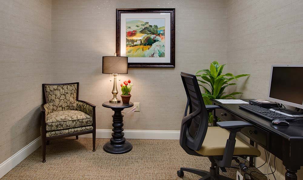 A cozy office corner with a comfortable chair, a small round table with a lamp and potted plant, an office chair, and framed artwork on the wall.