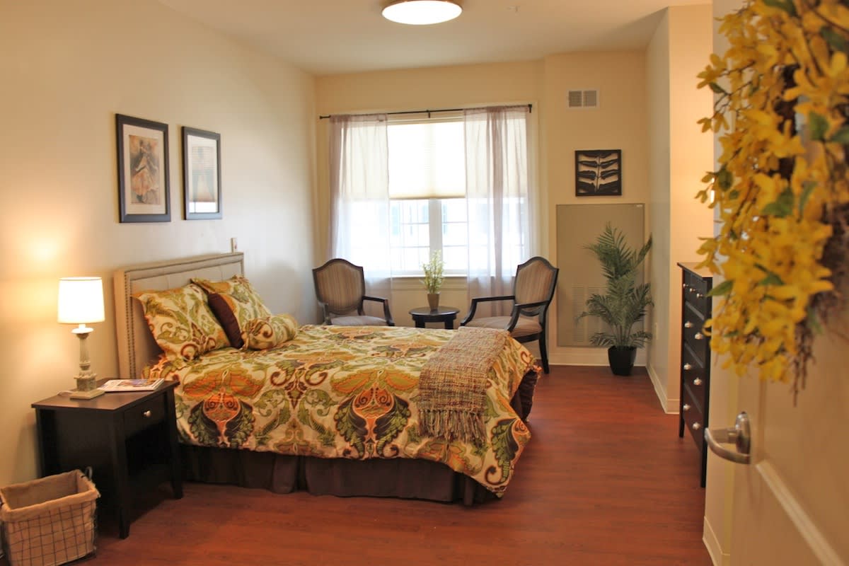 A cozy bedroom with elegant decor and wooden flooring, featuring a bed with patterned bedding, two chairs by the window, and framed artwork on the walls.