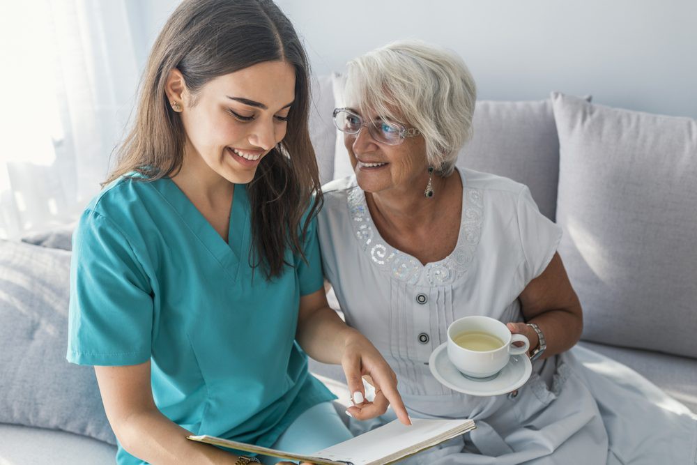 A nurse shares a light moment reading with an elderly woman enjoying a cup of tea.