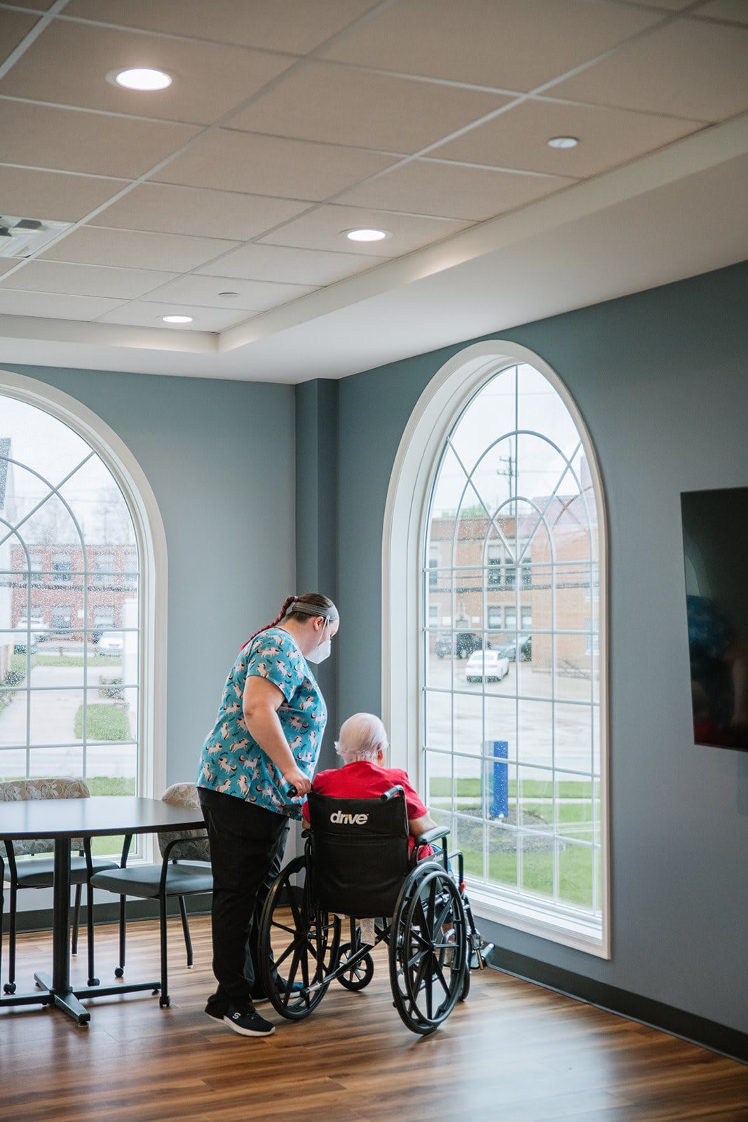 A caregiver assists an elderly person in a wheelchair by the window of a nursing home.