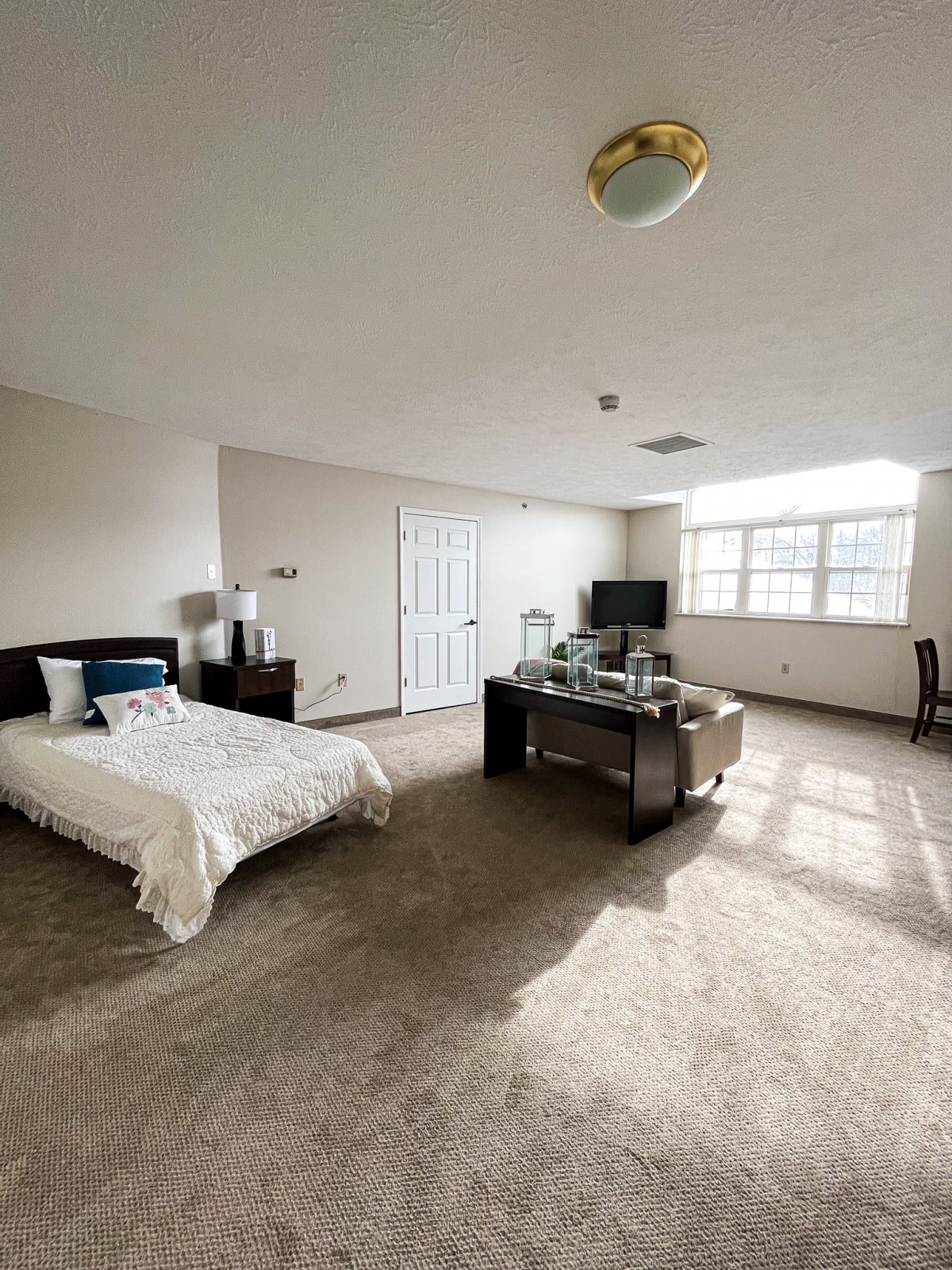 A spacious modern bedroom with a bed, desk, and large window providing ample natural light.