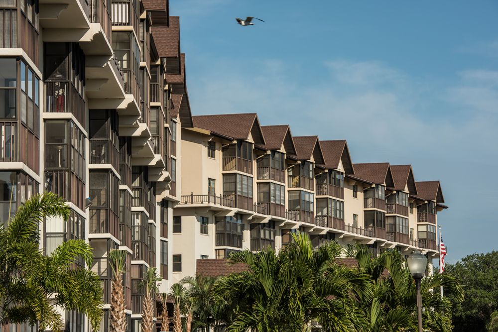A row of apartment buildings with balconies, under a clear blue sky, surrounded by palm trees.