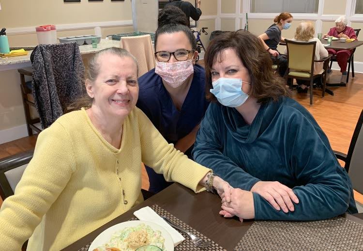 Three people gathered around a table, two wearing masks and smiling.