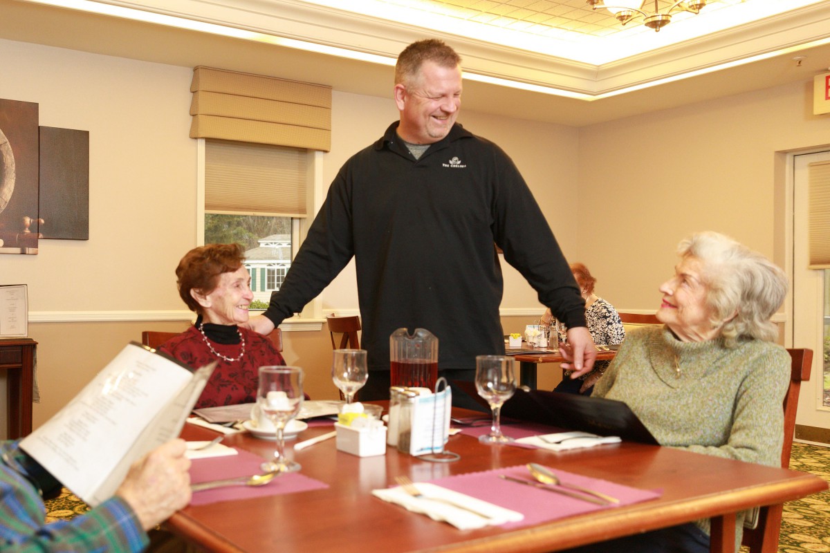 A man warmly interacts with elderly patrons at a dining table.