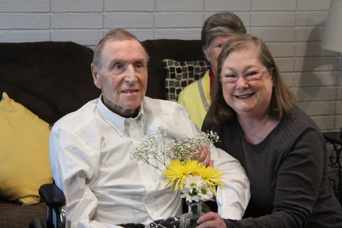 An elderly couple sitting together indoors, holding flowers and smiling.