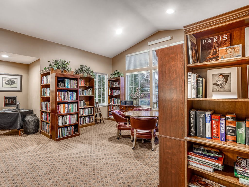 A cozy library room with bookshelves, a round table surrounded by chairs, and large windows letting in natural light.
