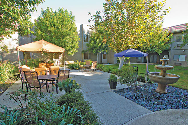 A serene garden with a patio area featuring outdoor furniture and gazebos, surrounded by lush trees and greenery.